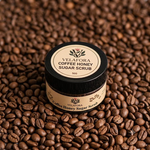A small jar of Velafora Coffee Honey Sugar Scrub sits on a bed of roasted coffee beans. The jar has a black lid with a beige label and a matching beige label wrapped around the body.