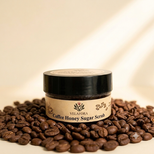 A jar of Velafora Coffee Honey Sugar Scrub sits on a bed of roasted coffee beans against a warm beige background with soft diagonal shadows.