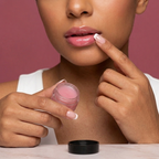 A close-up of a woman applying a glossy pink lip balm from a small clear jar with a black lid, set against a solid dusty rose background
