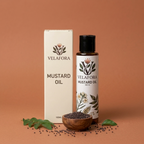 A bottle of Velafora mustard oil and its matching box are displayed on a warm terracotta background, styled with a small wooden bowl of mustard seeds and green leaves.