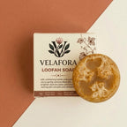 Velafora Loofah Soap bar placed in front of its cream-colored packaging box, set against a diagonally split terracotta and off-white background.