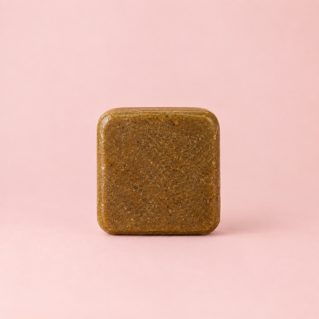 A square, rounded-corner bar of textured olive-brown soap or shampoo standing upright on a solid light pink background.