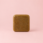 A square, rounded-corner bar of textured olive-brown soap or shampoo standing upright on a solid light pink background.