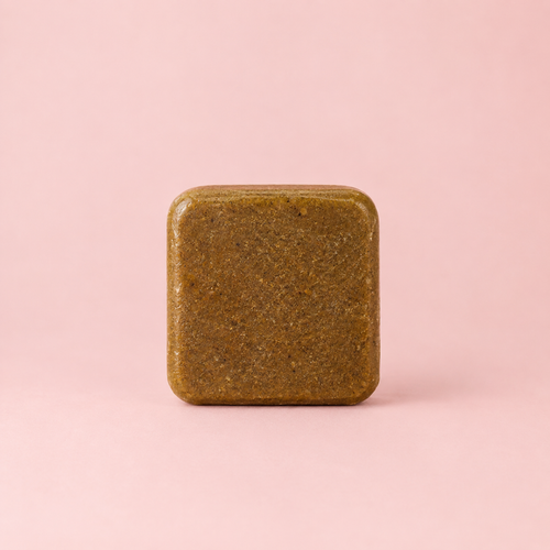 A square, rounded-corner bar of textured olive-brown soap or shampoo standing upright on a solid light pink background.
