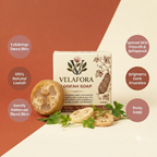 A boxed bar of Velafora Loofah Soap is centered on a split warm rust and cream background, with a sliced loofah and loose loofah pieces arranged in front. Text callouts around the product describe skin benefits: exfoliates dead skin, 100% natural loofah, gently removes dead skin, leaves skin smooth and refreshed, brightens dark knuckles, and is body soap.

